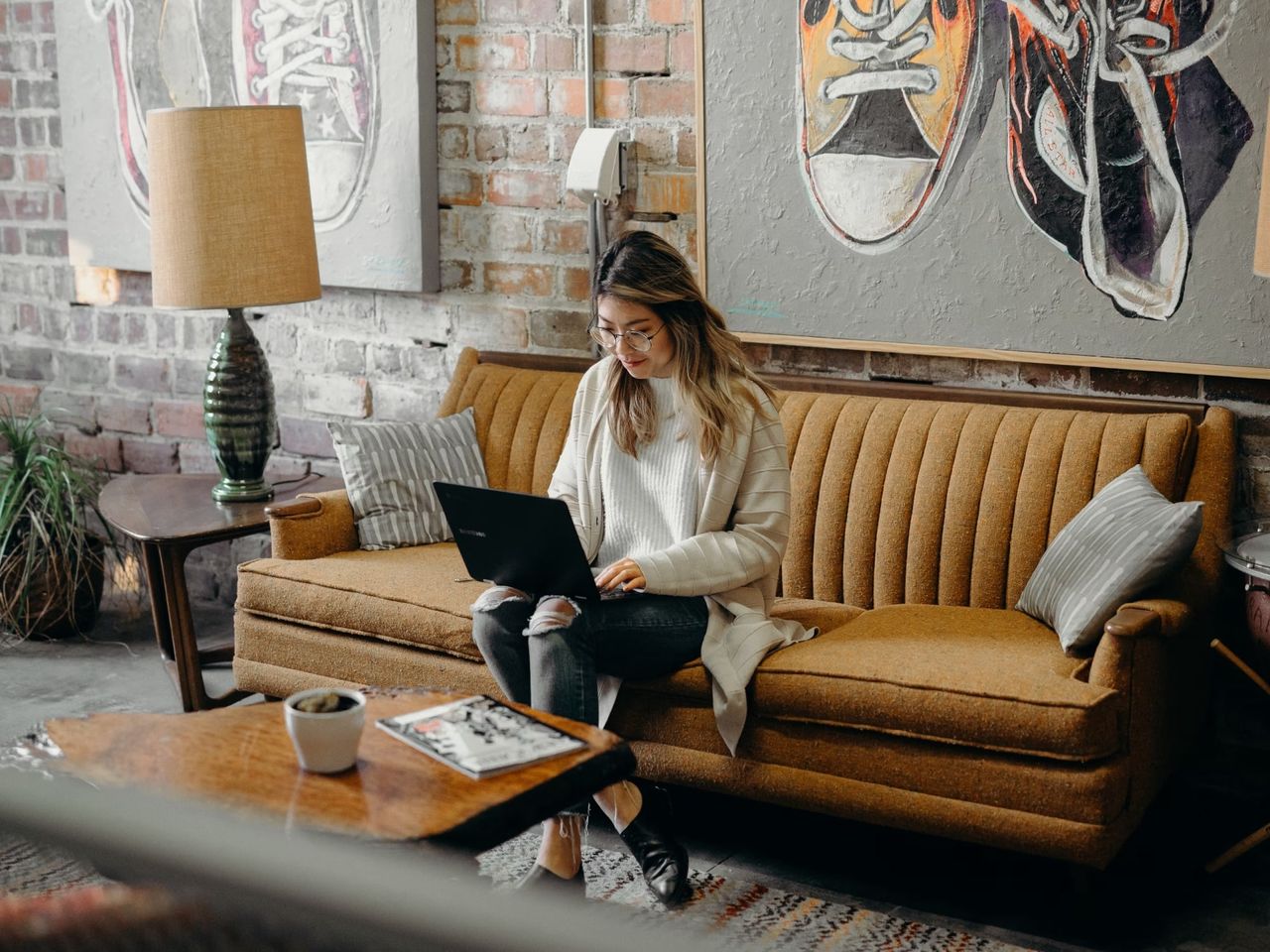 A woman on a vintage sofa typing on a laptop, with framed sneaker artwork on the wall behind her